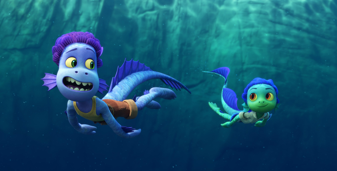 In a scene from Luca, Alberto (voiced by Jack Dylan Grazer) and Luca (voiced by Jacob Tremblay), two young sea monsters, swim in the ocean. Alberto, with light and dark blue scales and purple hair, wears brown shorts and a yellow tank top. Luca, who has teal and dark blue scales, wears blue shorts and a checkered green shirt. In the background, there is a rocky wall. 