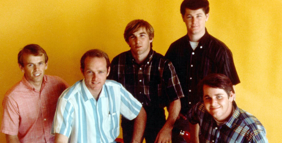 Members of the Beach Boys pose for a portrait against a yellow backdrop. From left to right are Al Jardine, Mike Love, Dennis Wilson, Brian Wilson, and Carl Wilson. They are all wearing short sleeve plaid shirts, pants and white loafers. Love and Carl Wilson are sitting on two red mopeds.