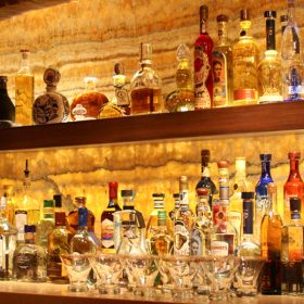 A well-stocked bar with a variety of liquor bottles displayed on illuminated shelves, set against a textured stone wall background.