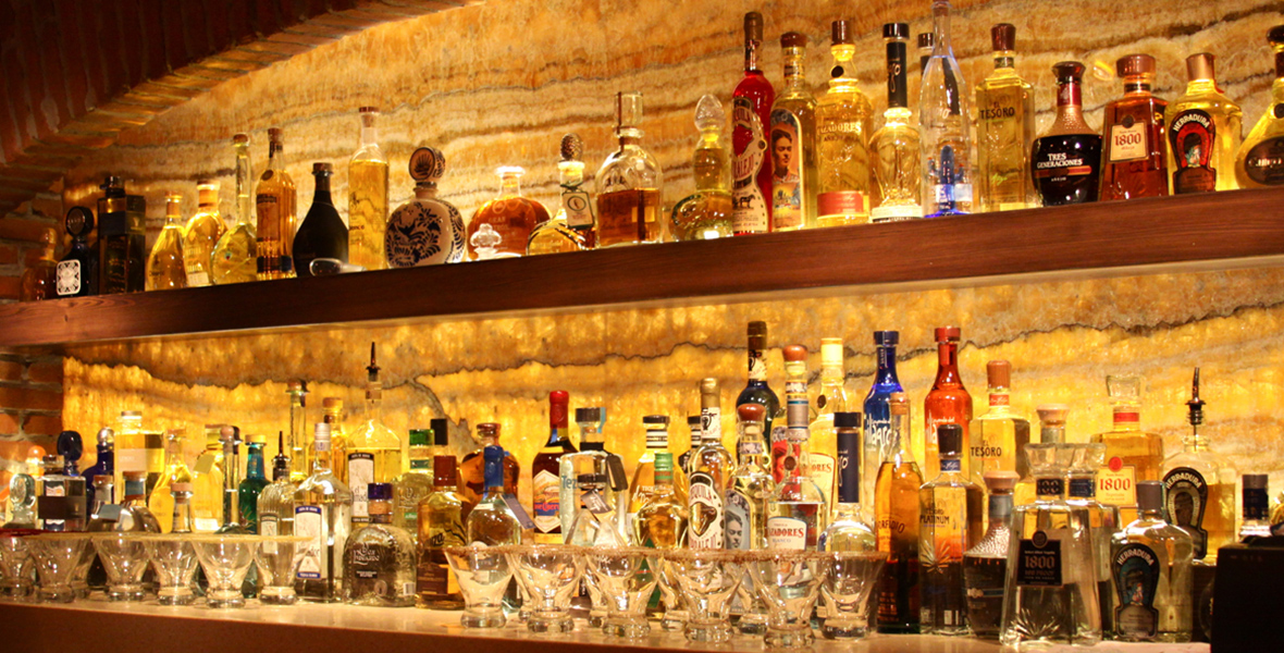 A well-stocked bar with a variety of liquor bottles displayed on illuminated shelves, set against a textured stone wall background.