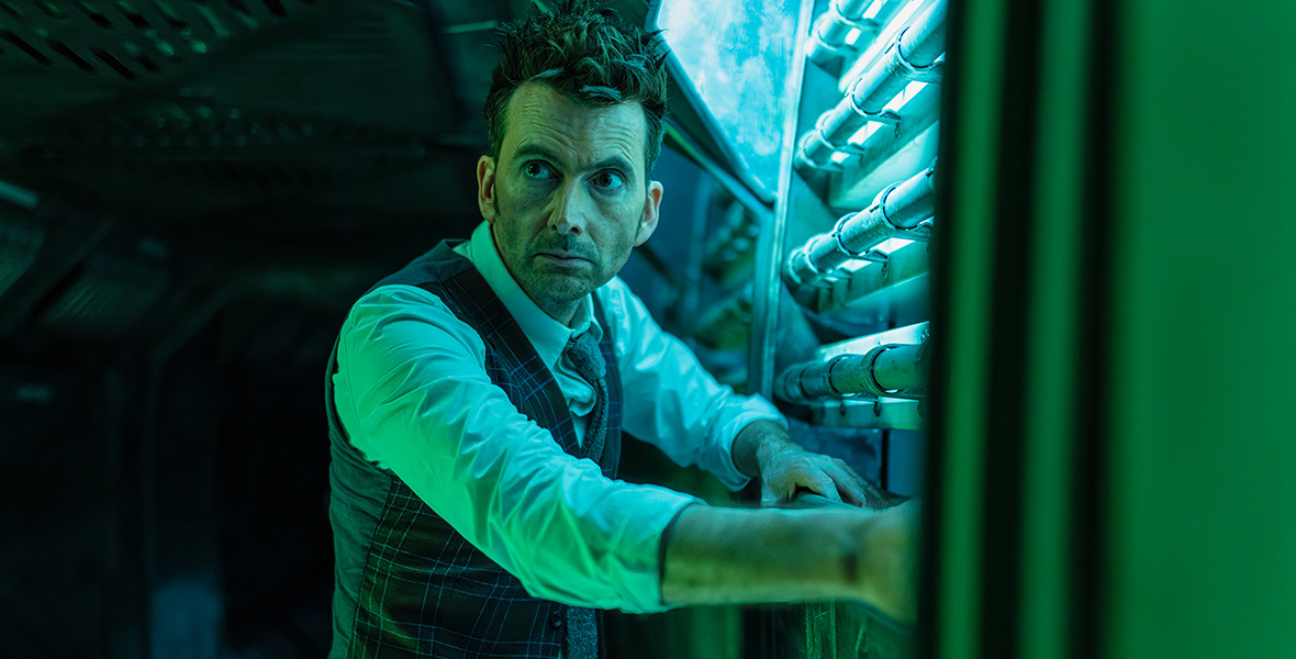 In an image from the Doctor Who special “Wild Blue Yonder” on Disney+, David Tennant as the Fourteenth Doctor is seen leaning against a wall of pipes; it’s dark and he’s looking off camera to the left, seriously. He’s wearing a tie and a plaid waistcoat and pants.