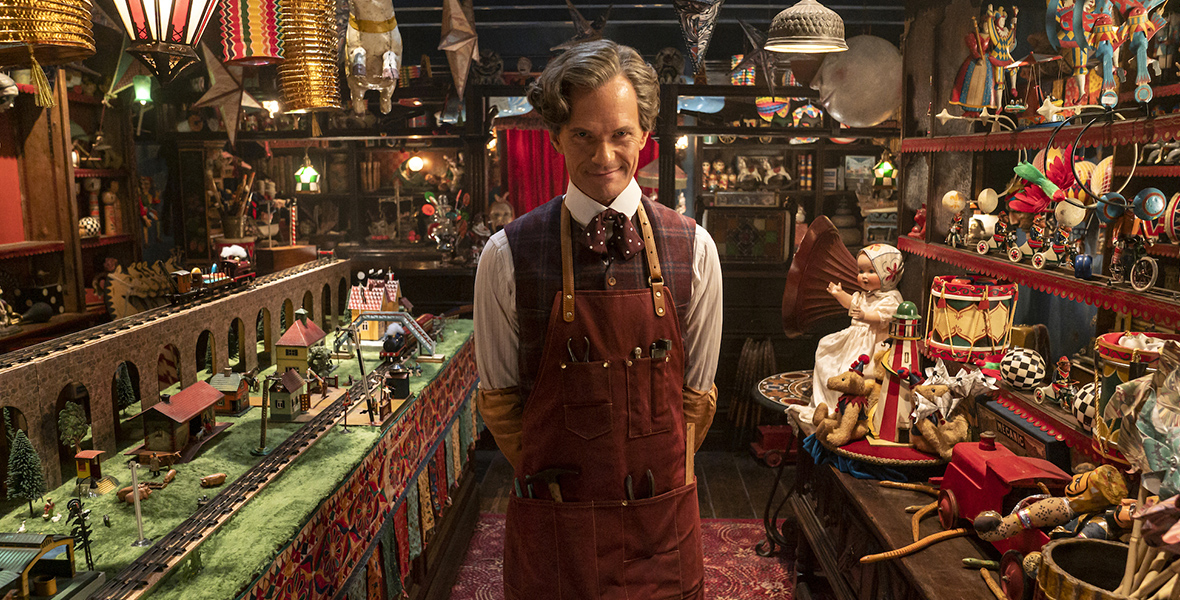 In an image from the Doctor Who special “The Giggle” on Disney+, Neil Patrick Harris as the Toymaker stands in a colorful toy shop. He is dressed in a brown apron, with pockets full of tools, and plaid waistcoat with a polka-dot bow tie. He’s wearing glasses, and is peering over them with a smirk. 