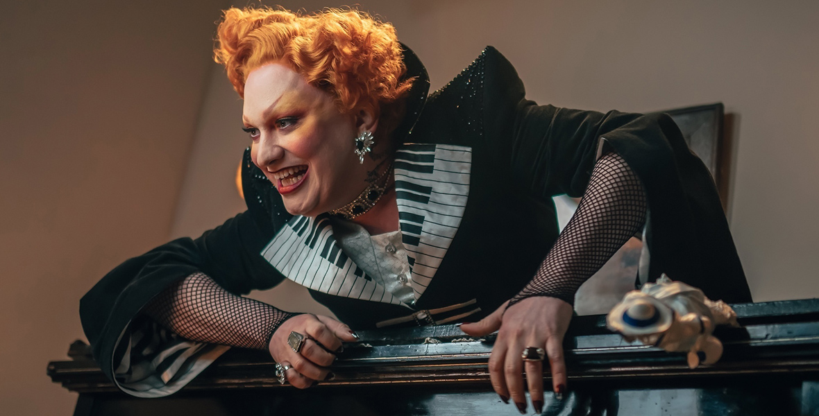 In an image from the Doctor Who episode “The Devil’s Chord” on Disney+, Jinkx Monsoon as Maestro is seen crawling out of an upright piano, menacingly. She has a shock of red hair as is wearing a sparkly black gown with piano keys around the neckline.