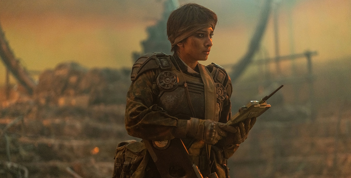 In an image from the Doctor Who episode “Boom” on Disney+, Varada Sethu as Mundy Flynn is seen wearing futuristic army-style fatigues and holding a communication tablet of some kind. Her head is bandaged, there’s a weapon slung around her toros, and she’s looking off to the right. Her surroundings look dark and post-apocalyptic. 