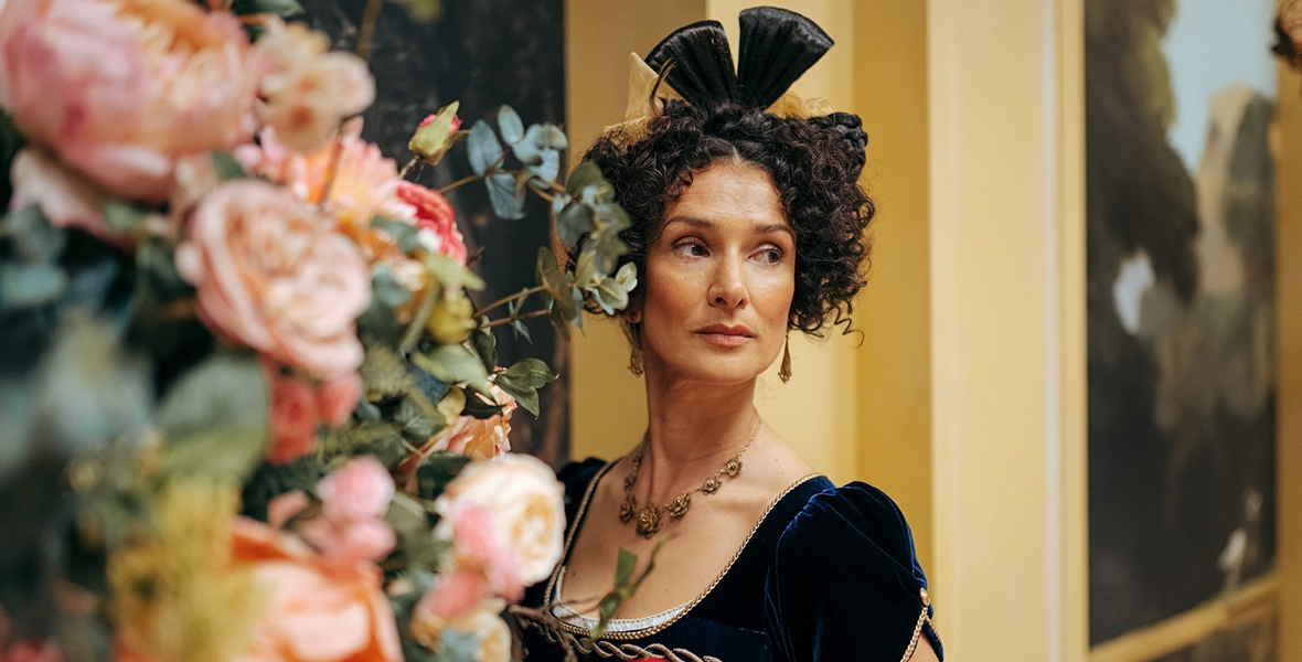 In an image from the Doctor Who episode “Rogue” on Disney+, Indira Varma as the Duchess of Pemberton is peering at someone to her right from behind a large display of pink flowers and stems. She is wearing an 1800s-style dress, gloves, and headpiece.