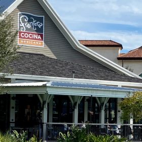 Exterior view of Frontera Cocina Mexicana, a Mexican restaurant, showcasing its distinctive gable-roofed building with a sign prominently displayed. The structure is surrounded by lush greenery, with outdoor seating visible on the wrap-around porch. The background includes additional buildings with Mediterranean-style architecture.