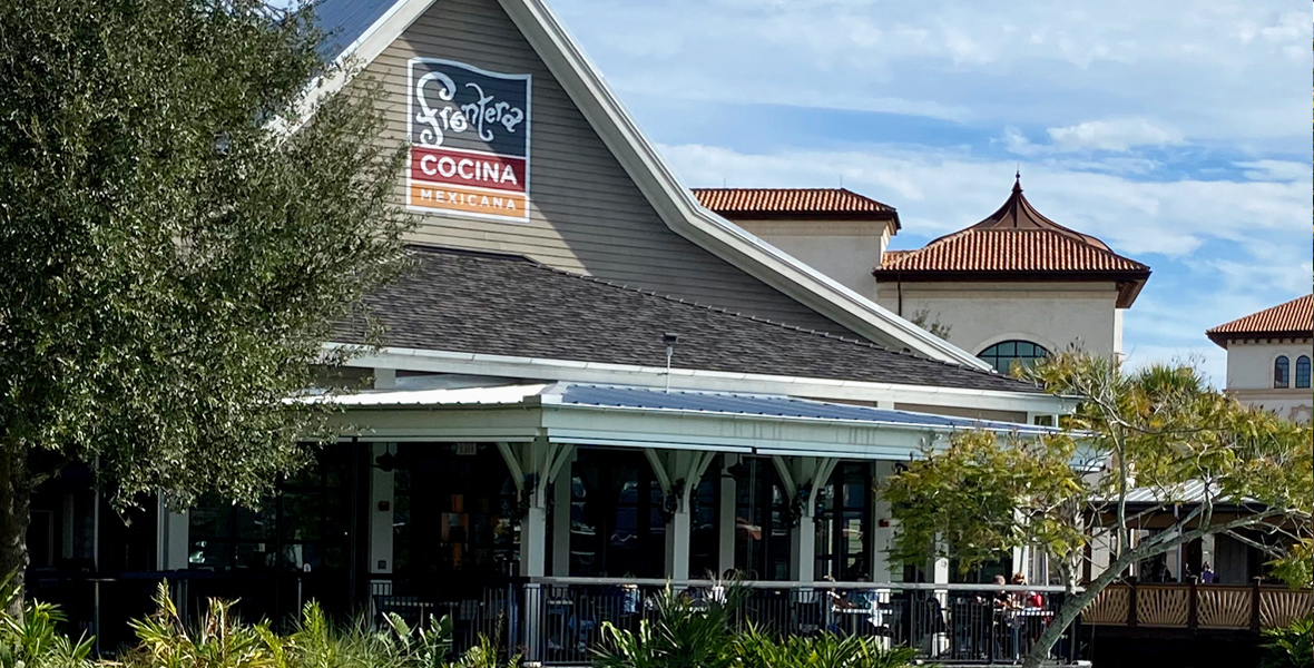 Exterior view of Frontera Cocina Mexicana, a Mexican restaurant, showcasing its distinctive gable-roofed building with a sign prominently displayed. The structure is surrounded by lush greenery, with outdoor seating visible on the wrap-around porch. The background includes additional buildings with Mediterranean-style architecture.