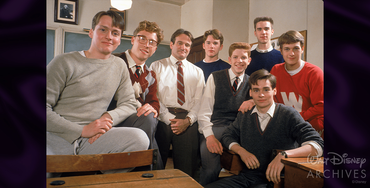The cast of Dead Poets Society poses in a classroom for a production photo. Gale Hansen as Charlie Dalton appears on the left, wearing grey sweatpants and a grey sweater, seated next to him, Allelon Ruggiero as Steven Meeks wears glasses and an argyle red sweater. Disney Legend Robin Williams as John Keating in the center middle is seen wearing a white-collar shirt with a red and blue striped tie. Ethan Hawke as Todd Anderson is next on the right, and wears a blue crewneck sweater, with Dylan Kussman as Richard Cameron next to him wearing a sleeveless V-neck dark gray sweater and a red and blue tie. To the right is James Waterston as Gerard Pitts who wears a blue crewneck sweater, with Josh Charles as Knox Overstreet on the far right, wearing a red sweater with a white “W” on the front. At the bottom right is Robert Sean Leonard as Neil Perry who wears a dark gray sweater and white-collared shirt that is unbuttoned at the collar.