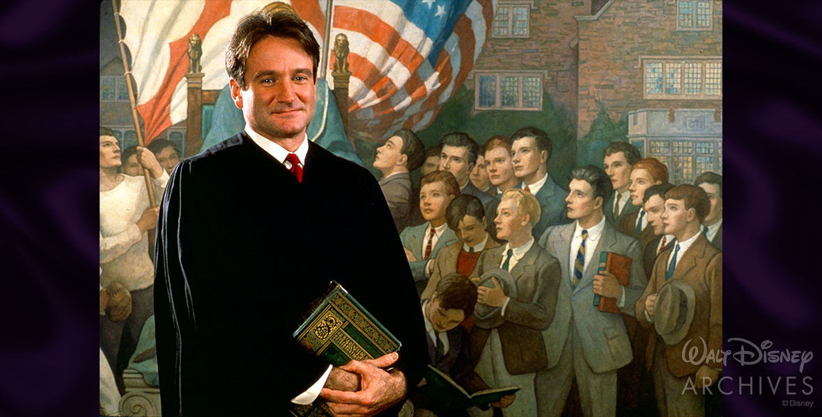 Disney Legend Robin Williams poses in front of a mural at St. Andrew’s School in Middletown, Delaware, the setting for Welton Academy, painted by N.C. Wyeth, that showcases schoolboys. Williams is dressed in classic academic black robes and a red tie and is holding a book by Shakespeare. 
