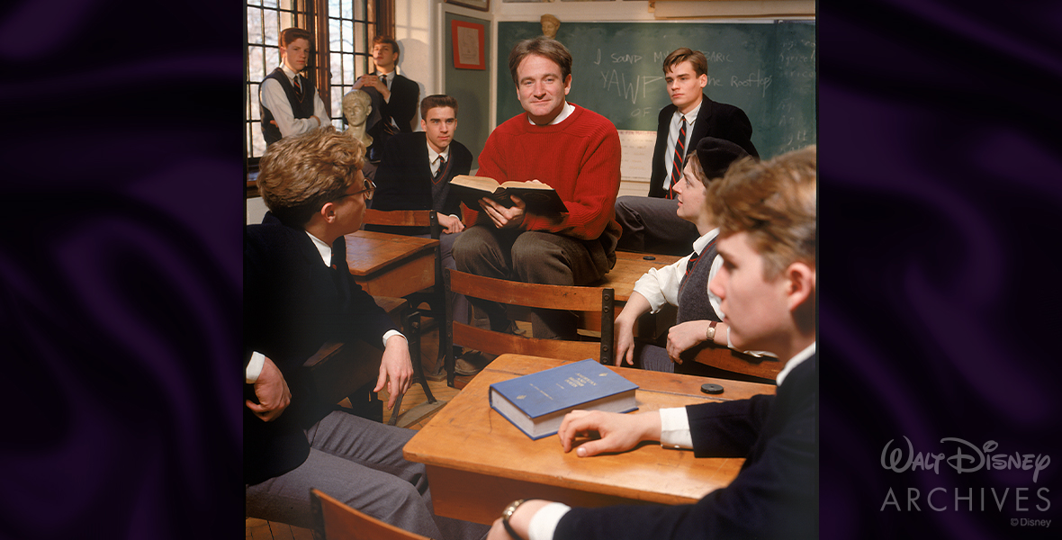 Disney Legend Robin Williams as John Keating poses for a production photo from Dead Poets Society, dressed in a red sweater and holding the book “Five Centuries of Verse” in the classroom setting. The seven main students sit around him, wearing their school uniforms