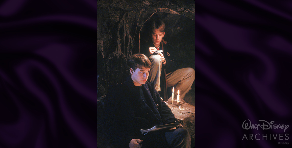 Josh Charles’ Knox Overstreet and Ethan Hawke’s Todd Anderson pose for a Dead Poets Society production photo while filming a scene inside the iconic cave. Charles on the bottom left wears a dark navy trench coat and looks forward, while Hawke wears khaki slacks and a dark navy trench coat and is seen writing in a small notebook. On a rock between the two are two lit candles. 