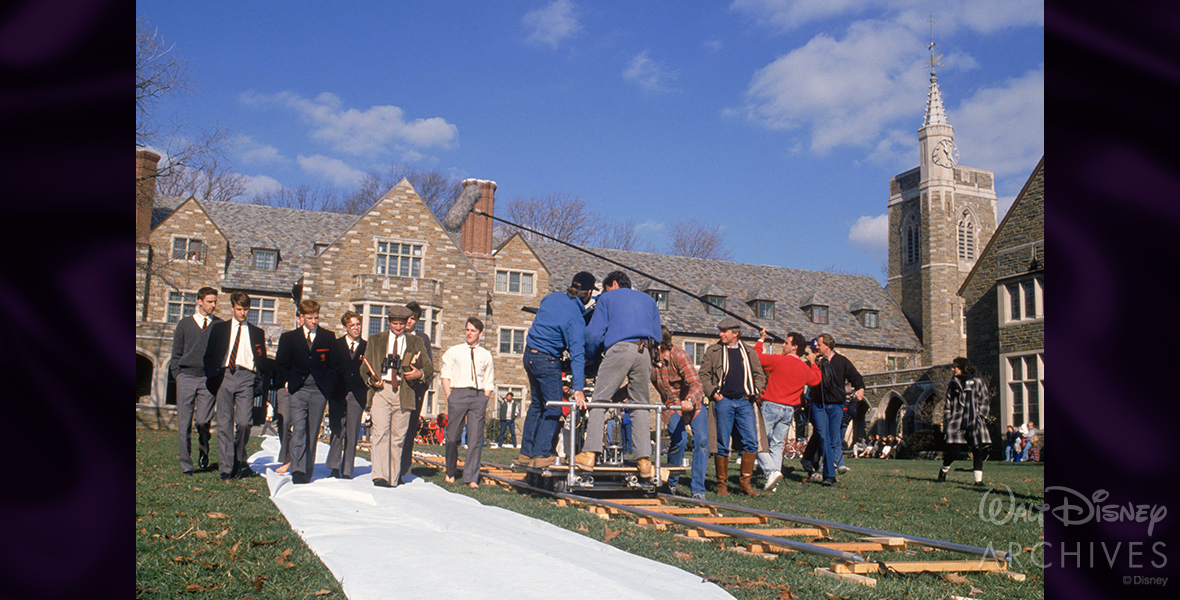 A behind-the-scenes photo from Dead Poets Society, outside at St. Andrew’s School on a sunny fall day, showcasing the camera crew and actors as they walk alongside Disney Legend Robin Williams while filming an outdoor scene. The boys are in their school uniforms and Williams is dressed in slacks, a white shirt, and a tie with a green suit jacket. 