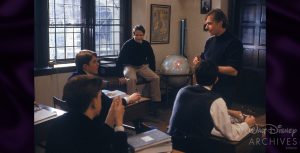 A Dead Poets Society behind-the-scenes photo of director Peter Weir (right) addressing a young Ethan Hawke (left), who plays Todd Anderson, and Robin Williams’ John Keating, sitting on a table next to a globe (middle). They are all in the classroom with a window on the left.