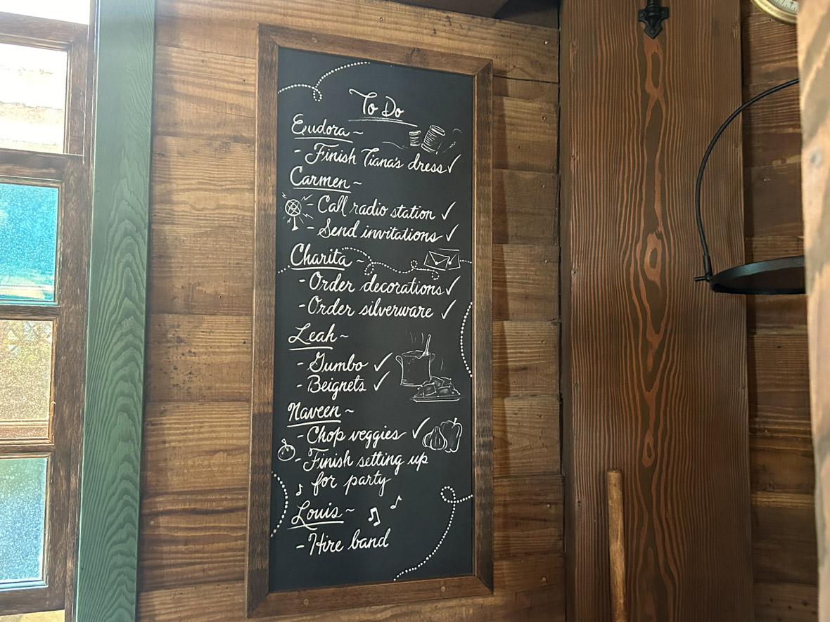 A to-do list written in white chalk on a black chalk board, featuring tasks that characters from The Princess and the Frog need to or have completed before the big Mardi Gras party. Included on the list with the character names are the names Carmen, Charita, and Leah.