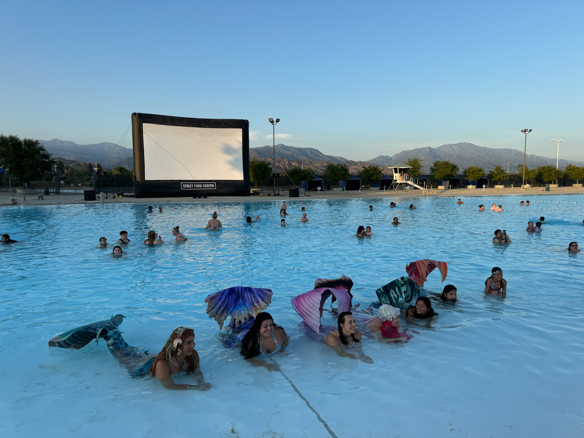 People enjoy a refreshing swim in a large outdoor pool during a movie screening event. The scene is set against a backdrop of mountains, with an inflatable movie screen showing the logo of Street Food Cinema. Several attendees, including some dressed as mermaids with colorful tails, relax and float in the water, preparing to watch a film as the sun sets.