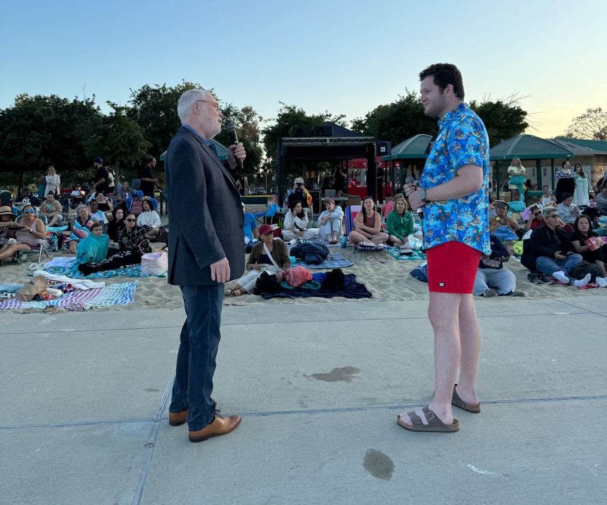  Two men stand facing each other in conversation at an outdoor event. One man, wearing a blue blazer and jeans, holds a microphone while the other man, dressed in a blue Hawaiian shirt and red shorts, listens. The background features a crowd of people sitting on blankets, enjoying a beachside gathering.