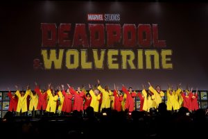A massive choir wearing red and yellow robes sings onstage at San Diego Comic-Con. The Deadpool & Wolverine logo is displayed on a giant screen behind them.