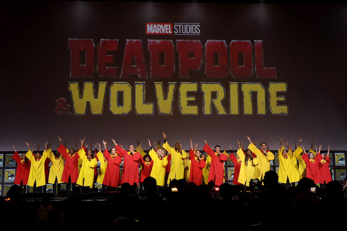 A massive choir wearing red and yellow robes sings onstage at San Diego Comic-Con. The Deadpool & Wolverine logo is displayed on a giant screen behind them.