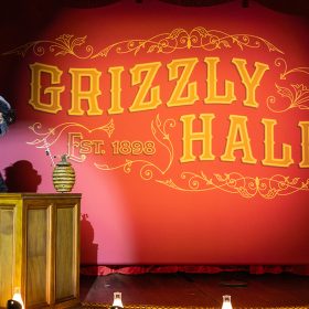In an image from Walt Disney World’s Country Bear Musical Jamboree, on a vaudeville style stage is a tall grizzly bear seated at a small upright piano on the left. The bear is Gomer, and he wears a white collar with a red-and-blue-striped tie and a lavender hat. Atop the piano is a round beehive with two straws sticking out of the to. At the back of the stage is a red curtain with writing on it that says “Grizzly Hall, Established 1898.” Two spotlights illuminate Gomer and the curtain.
