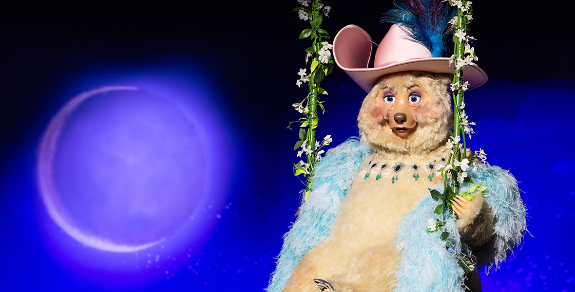 In an image from Walt Disney World’s Country Bear Musical Jamboree, a plump blond bear with a sky-blue boa and a pink cowgirl hat (adorned with a few tall blue-and-purple peacock feathers) is seated on a swing made of thick and leafy vines, hanging above an unseen stage. This is the bear singer Teddi Barra, who has blue eyes and pink cheeks and wears a necklace made of emerald and onyx-colored gems. Behind Teddi is a backdrop painted deep blue with a large crescent moon on the left side.