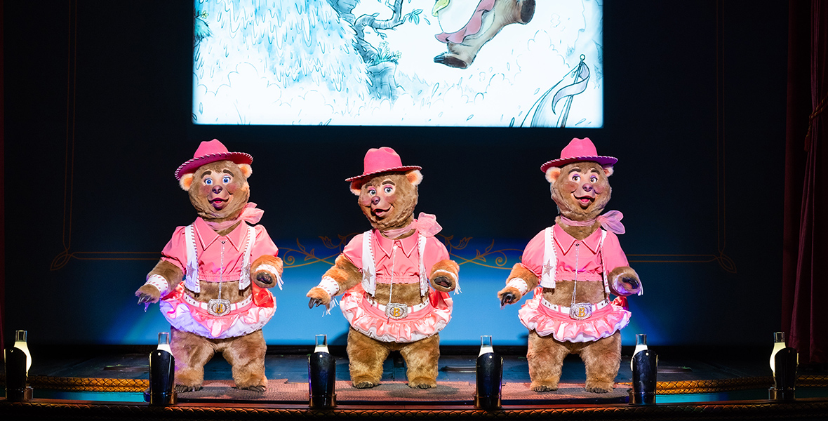 In an image from Walt Disney World’s Country Bear Musical Jamboree, and seen standing in a row on a vaudeville-style stage, are three tan-furred bears wearing identical outfits of pink crop-top blouses, pink-and-white skirts with white belts and bejeweled buckles, white vests with big pink stars on each side, and pink ranger-style hats with white trim. These are Bunny, Bubbles, and Beulah, the triplets, and they all wear blue eye shadow and pink lace scarves tied with a bow around their necks. Behind them is a video screen that appears to depict one of them in danger, hanging precariously from a thin tree that leans out over a cascading waterfall, the bottom of which is not visible.