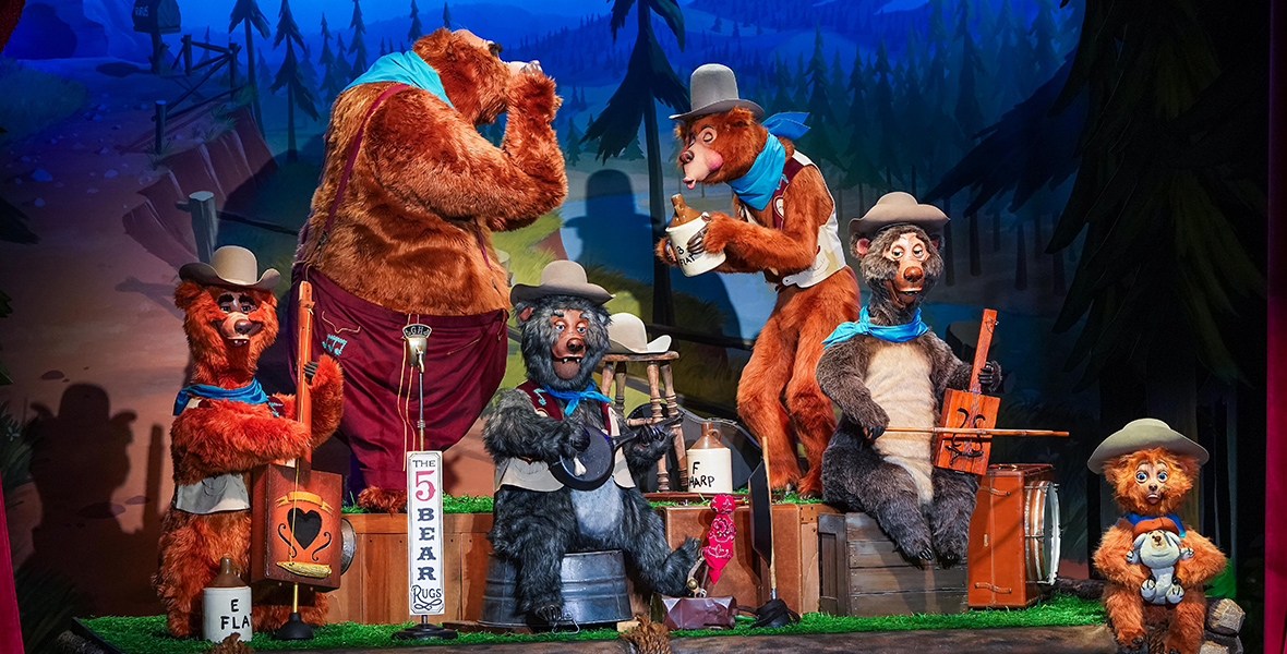 In an image from Walt Disney World’s Country Bear Musical Jamboree, and seen on a log platform atop a vaudeville-style stage, is a band made up of five bears. From left to right we see a short reddish bear playing a homemade standup bass with just one string; a huge brown bear playing a harmonica; a small black bear playing a homemade banjo; a tall thin bear whose pursed lips are blowing into a jug; and a medium-sized black bear with a tan chest and stomach seated on a box and playing a homemade fiddle. All the bears have cowboy hats on their heads except the harmonica player, whose hat sits on a wooden stool in front of him. All the bears have blue neck scarves; three of them have vests; and the harmonica player wears pants. In front of the harmonica player is an old-fashioned microphone with a sign attached to the microphone stand reading, “The 5 Bear Rugs.” A sixth bear, a pouting reddish cub, sits on the right front corner of the log platform, wearing a cowboy hat and holding a stuffed animal toy. The backdrop behind the bears depicts a forested mountain scene in cartoon style.