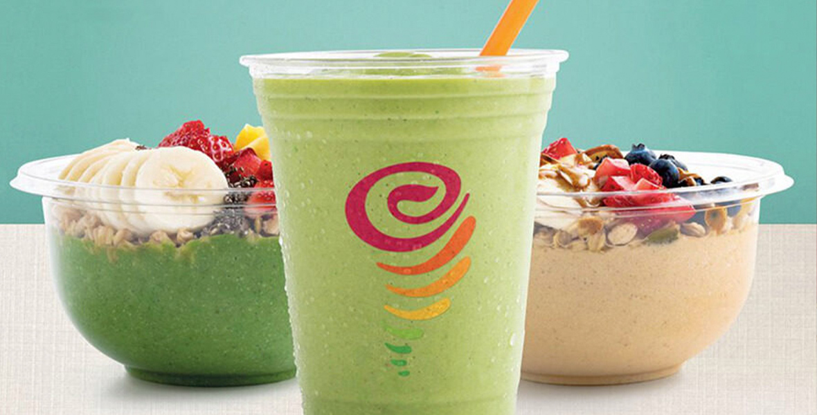 The image shows a refreshing green smoothie in a clear plastic cup with an orange straw. The cup features the Jamba logo, a colorful swirl design. Behind the smoothie are two smoothie bowls in clear bowls. The bowl on the left contains a green smoothie base topped with banana slices, strawberries, blueberries, and granola. The bowl on the right has a creamy beige base topped with strawberries, blueberries, and granola drizzled with a sauce.