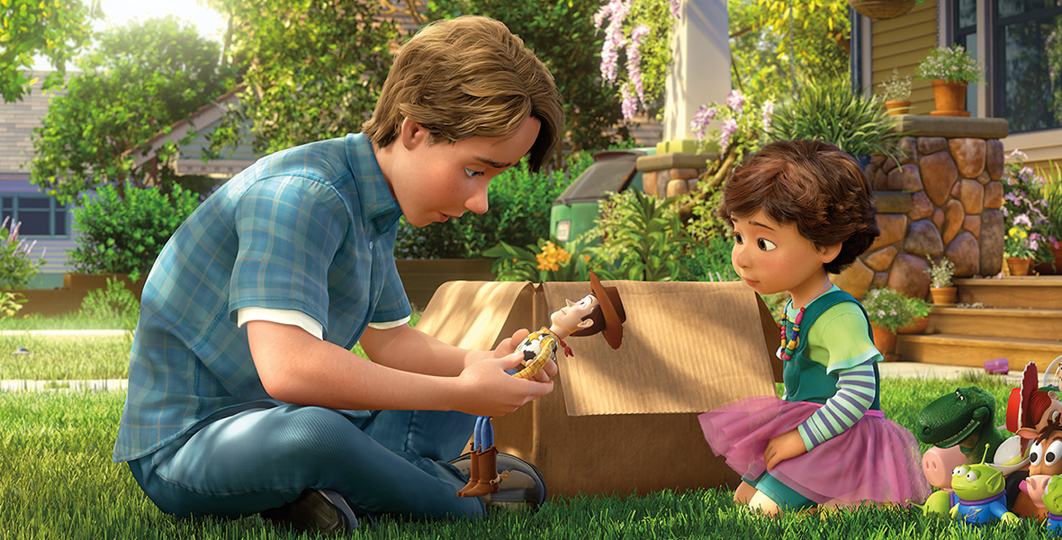 In an image from Toy Story 3, Andy sits in the grass and holds Woody in both hands. Bonnie kneels opposite Andy, in front of the other toys he’s given to her. A cardboard box is opened behind them.