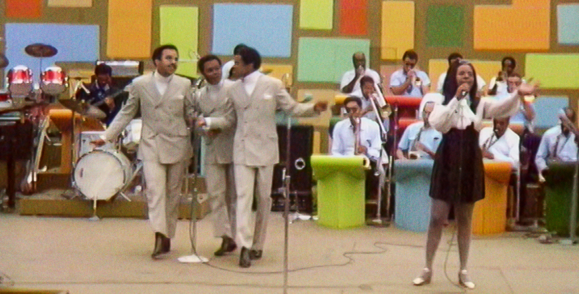 In an image from Summer of Soul (...Or, When the Revolution Could Not Be Televised), Gladys Knight & the Pips are seen performing on stage during the Harlem Cultural Festival in 1969. A band made up of around 15 musicians is seen behind them, and a colorful wall is behind the entire group; the word “Festival” can be seen in large letters, in white, on said wall.