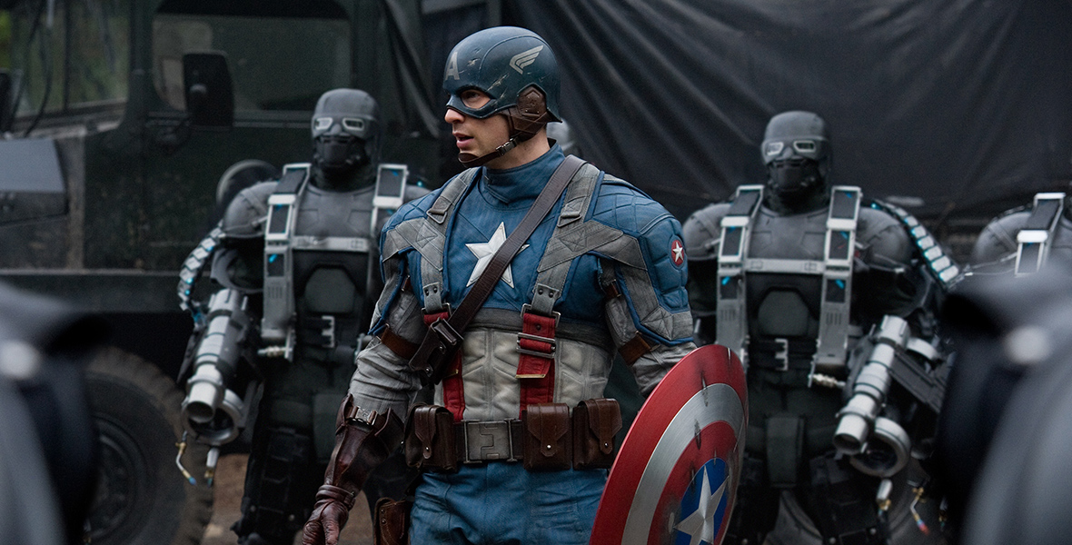 In an image from Captain America: The First Avenger, Chris Evans as seen as the title character, wearing his traditional outfit and carrying his shield. He is surrounded by several other heavily fortified humans, and there is a military truck in the background.