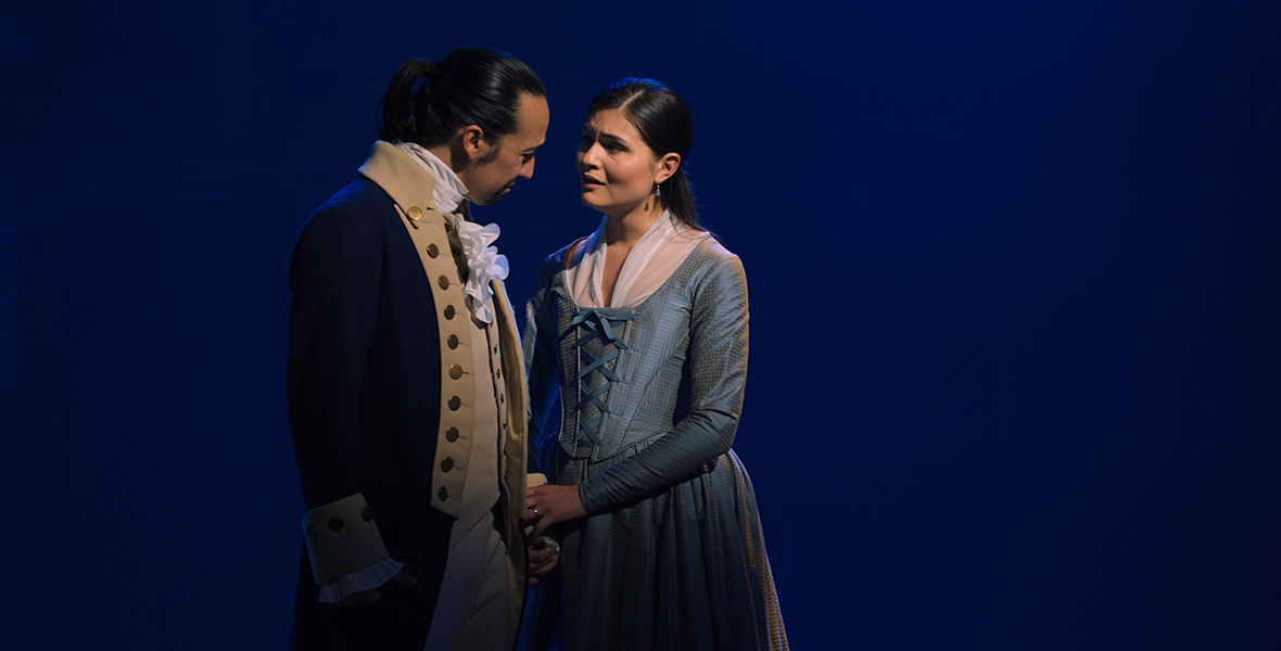 In an image from the Broadway production of Hamilton, filmed for streaming on Disney+, Lin-Manuel Miranda and Philippa Soo are seen as Alexander and Eliza Hamilton, respectively. They are standing together on a dark stage; Soo is singing to Miranda and holding his hand.