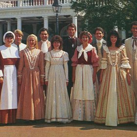 The Voices of Liberty a capella vocal group stands in two lines, shoulder to shoulder, in front of the American Adventure pavilion at EPCOT in the 1980s. In the front row are seven women, while eight men stand behind them. All are dressed in American Colonial costumes, with ankle-length dresses and a few aprons and caps on the women and knee-length socks worn below loose pants gathered below the knee on the men, who also sport ascots, vests, and jackets.