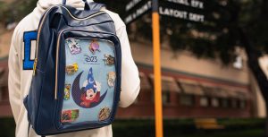 The image shows a person wearing a blue backpack adorned with various Disney-themed pins. The backpack features a prominent design of Mickey Mouse in his iconic Sorcerer's Apprentice costume from "Fantasia," with the D23 logo and the phrase "The Official Disney Fan Club" underneath. The pins on the backpack include characters and symbols from different Disney movies and themes, including a D23 Gold Member pin. The person is standing outdoors, with a signpost in the background that has blurred text indicating it might be a location within a Disney park or studio.