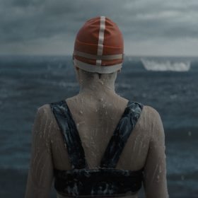 Daisy Ridley, as Trudy Ederle in Disney’s Young Woman and the Sea, is seen with her back toward the camera, wearing a red bathing cap and the top of a black two-piece bathing suit, with a grease-like substance applied on her body. A dark ocean lays before her and the sky is overcast with clouds and coming darkness.
