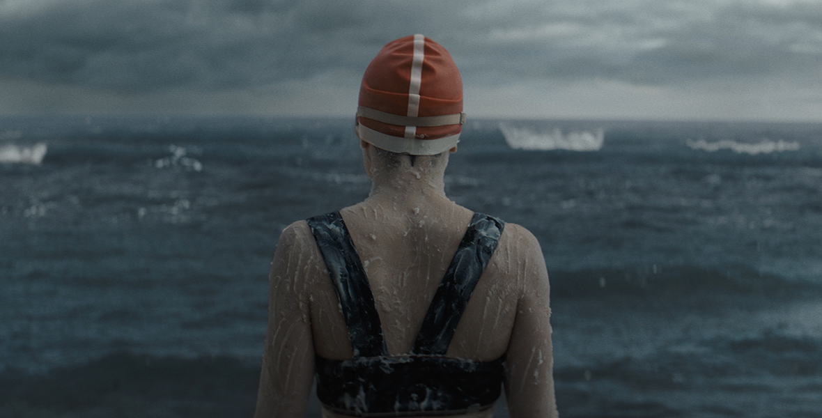 Daisy Ridley, as Trudy Ederle in Disney’s Young Woman and the Sea, is seen with her back toward the camera, wearing a red bathing cap and the top of a black two-piece bathing suit, with a grease-like substance applied on her body. A dark ocean lays before her and the sky is overcast with clouds and coming darkness.