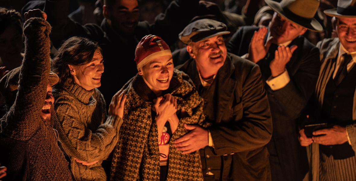 L-R, Tilda Cobham-Hervey as Meg Ederle, Daisy Ridley as Trudy Ederle, and Kim Bodnia as Henry Ederle Sr. stand on a beach at night illuminated in the cast of bonfires with a celebratory crowd around them, in Disney’s Young Woman and the Sea.