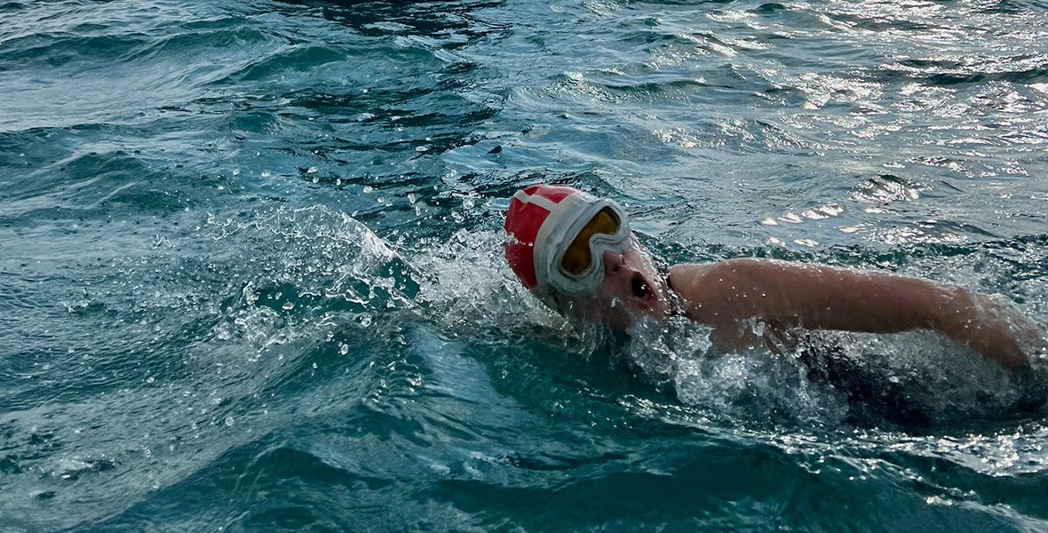 Daisy Ridley is seen swimming in open water during production of Disney’s Young Woman and the Sea. She wears a red bathing cap and large white googles with her face and right arm turned toward the camera. 