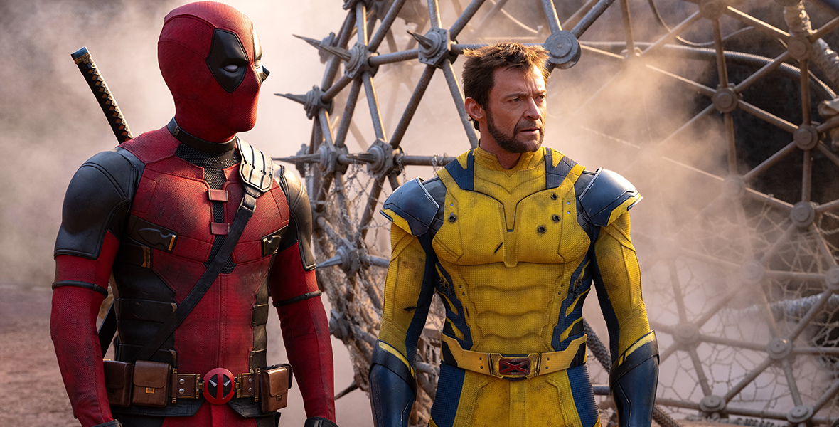 In a scene from Marvel Studios’ Deadpool & Wolverine, a masked Deadpool (left) wears a red and black suit next to Wolverine (right) who wears a blue and yellow suit that is scuffed and has several bullet holes in it. They are standing in a postapocalyptic landscape.