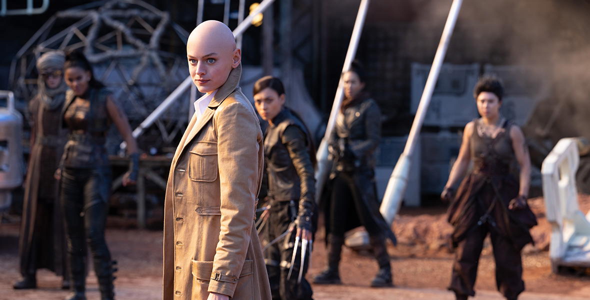 In a scene from Marvel Studios’ Deadpool & Wolverine, a bald Cassandra Nova wears a camel trench coat with a popped collar and stands in a postapocalyptic landscape. She is surrounded by several mutants, all of whom appear slightly out of focus.
