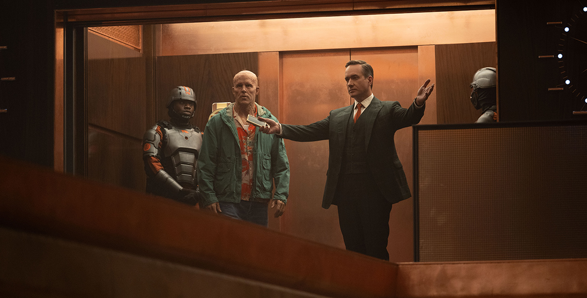 In a scene from Marvel Studios’ Deadpool & Wolverine, a TVA agent stands behind Wade Wilson (center) and Mr. Paradox (right), who is showing off TVA headquarters. The TVA agent is wearing an orange and black uniform; Wade is wearing a green jacket, an orange Hawaiian print shirt, and jeans; and Mr. Paradox is wearing a suit.