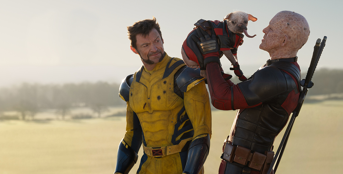 In a scene from Marvel Studios’ Deadpool & Wolverine, Wolverine wears a blue and yellow suit. He grimaces as an unmasked Deadpool, wearing a red and black suit, lifts Dogpool in the air. Dogpool is wearing a red and black suit and her tongue is sticking out.