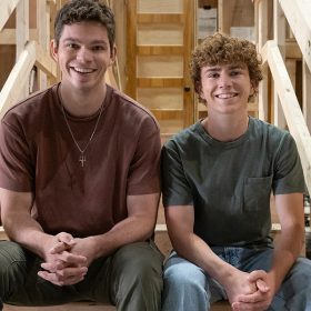Daniel Diemer (left) and Walker Scobell (right) pose for a photo on the set of Percy Jackson and the Olympians. They are smiling and sitting on wooden staircase. In a nod to his role as Poseidon's son, Diemer is wearing a necklace featuring a trident pendant.