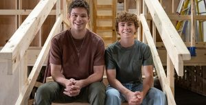 Daniel Diemer (left) and Walker Scobell (right) pose for a photo on the set of Percy Jackson and the Olympians. They are smiling and sitting on wooden staircase. In a nod to his role as Poseidon's son, Diemer is wearing a necklace featuring a trident pendant.