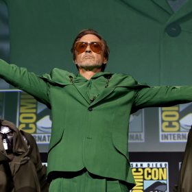 Robert Downey, wearing an all green suit, extends both arms wide. He is holding a Doctor Doom mask in his right hand and is surrounded by other robed figures wearing Doctor Doom masks.