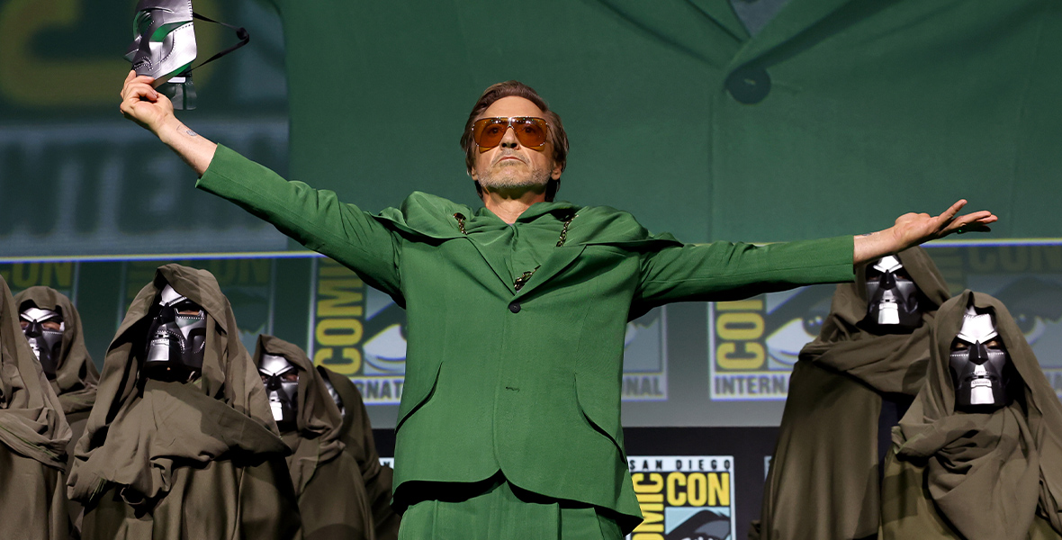 Robert Downey, wearing an all green suit, extends both arms wide. He is holding a Doctor Doom mask in his right hand and is surrounded by other robed figures wearing Doctor Doom masks.