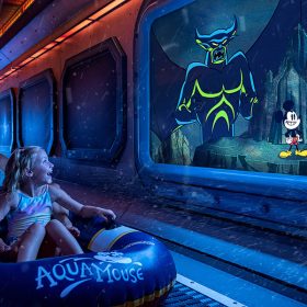 An adult and a child ride an inflatable tube on the AquaMouse. They are looking at an animated scene on a screen depicting Chernabog, Mickey Mouse, and Minnie Mouse