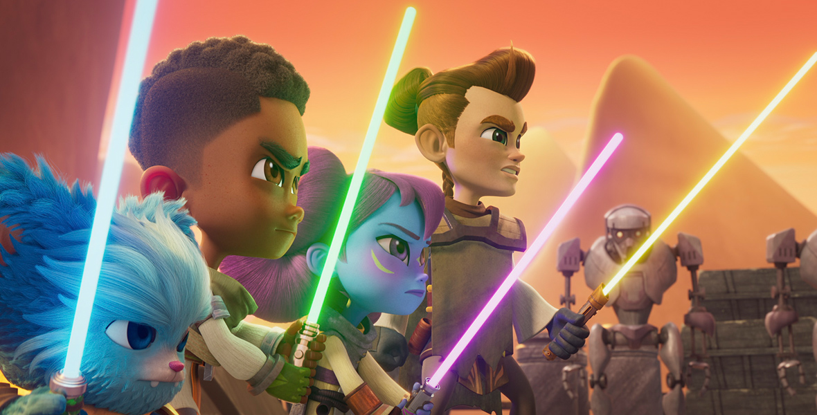 In a scene from Star Wars: Young Jedi Adventures, Nubs (voiced by Dee Bradley Baker), a blue-furred bear-like creature, is holding a blue lightsaber, ready to attack. Similarly, from left to right, Kai Brightstar (voiced by JeCobi Swain) holds a green lightsaber, Lys Solay (voiced by Juliet Donenfeld) holds a pink lightsaber, and Wes Vinik (voiced by Gunnar Sizemore) holds a yellow lightsaber. In the bottom right of the frame are three brown robots. The background features a desert-like setting, with red, orange, and yellow hues and two yellow mountains.