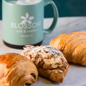 The image features three delicious-looking pastries arranged on a white rectangular plate. From left to right, there is a chocolate croissant, an almond croissant topped with sliced almonds and powdered sugar, and a classic croissant. In the background, there is a light green coffee mug with the logo of "Blossom Café & Market, Anaheim, CA" printed on it.