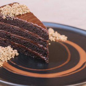 The image shows a slice of rich, layered chocolate cake on a black plate. The cake has several moist layers of chocolate sponge, separated by thick, glossy chocolate ganache. The top of the cake is garnished with a sprinkle of crunchy, crumbly topping. The plate is decorated with a swirl of chocolate sauce, adding an artistic touch to the presentation.