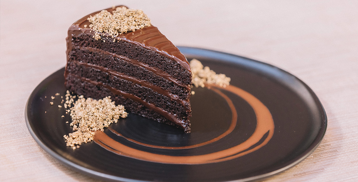 The image shows a slice of rich, layered chocolate cake on a black plate. The cake has several moist layers of chocolate sponge, separated by thick, glossy chocolate ganache. The top of the cake is garnished with a sprinkle of crunchy, crumbly topping. The plate is decorated with a swirl of chocolate sauce, adding an artistic touch to the presentation.